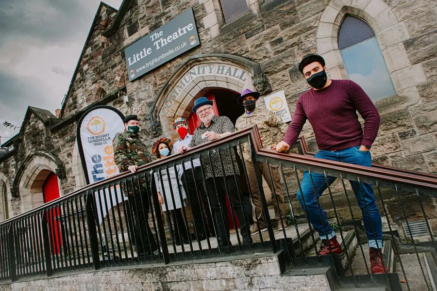 A man in a burgundy jumper stands on exterior steps of a stone theatre building with a group of people wearing masks on an upper balcony.