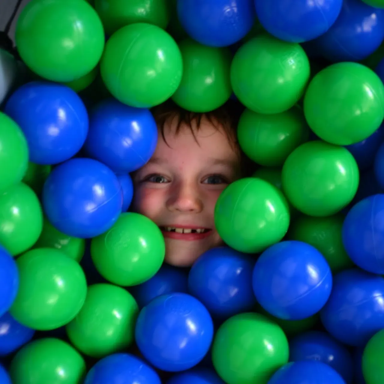Child surrounded by green and blue plastic balls in a ball pit, smiling at the camera.