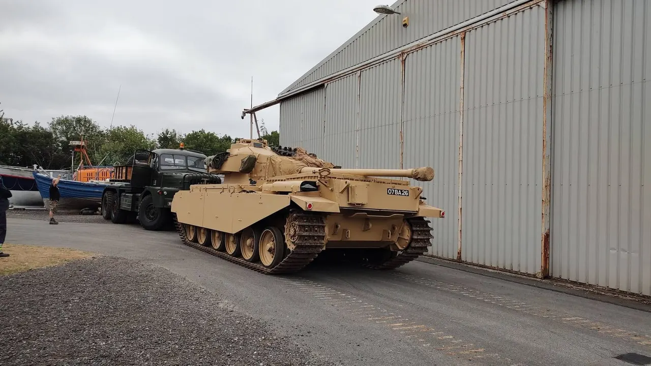 A tan military tank being transported on a lorry outside a corrugated metal hangar.