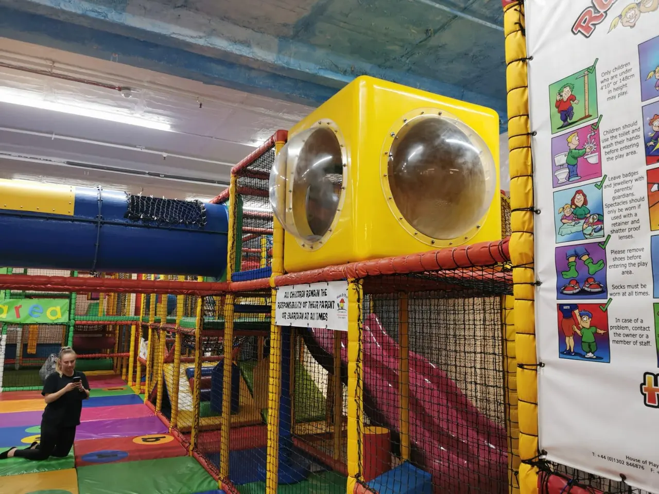 Child playing in an indoor soft play area with colourful padded equipment and a yellow structure with round windows.
