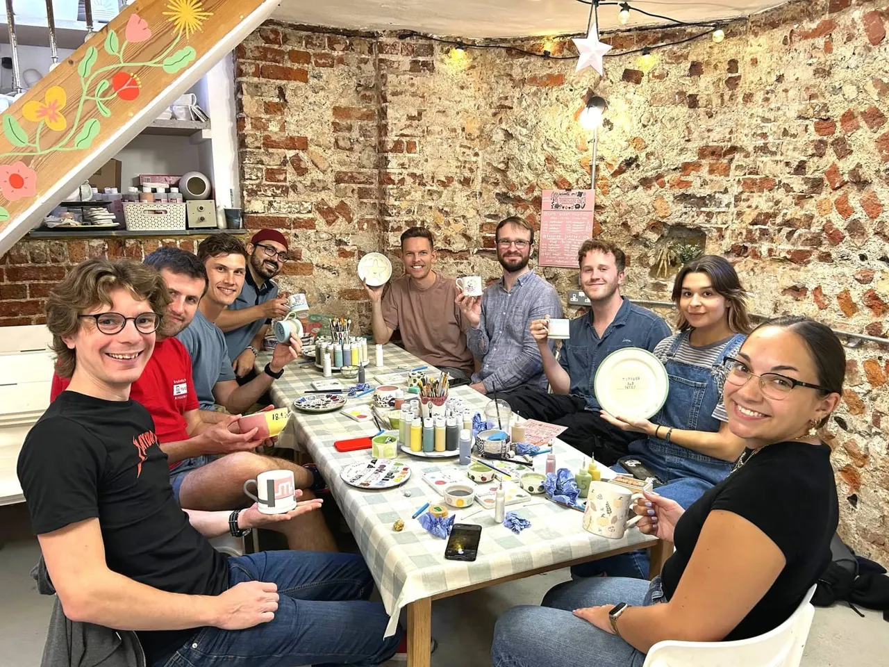 A group of nine people sit around a table at a pottery painting session, holding decorated plates and cups, smiling at the camera.