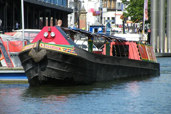 Red and black narrowboat moored on a city canal with urban buildings in the background.