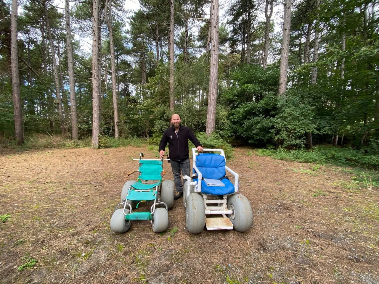 Man standing between two all-terrain buggies in a sandy woodland clearing surrounded by tall trees.