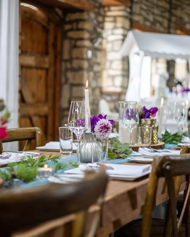 Elegant dining table set with purple flowers, candles, glassware and place settings in a stone-walled venue.