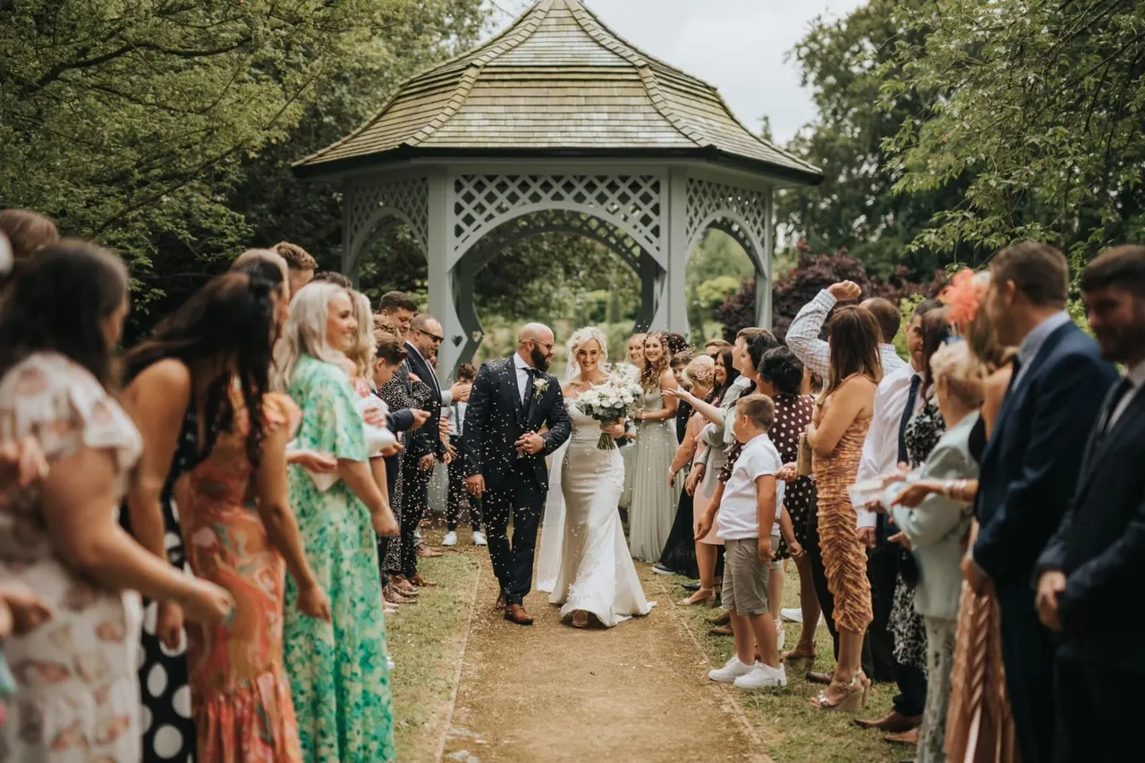 Bride and groom walk through garden with confetti as wedding guests cheer beside a decorative wooden gazebo.