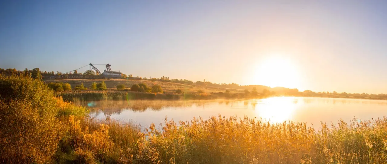 Sunset view across a lake with industrial mining structures on the far bank, surrounded by autumn vegetation and trees.