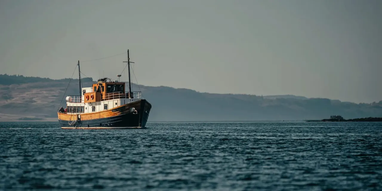An orange and black vintage passenger vessel moored on calm water with forested hills in the background.
