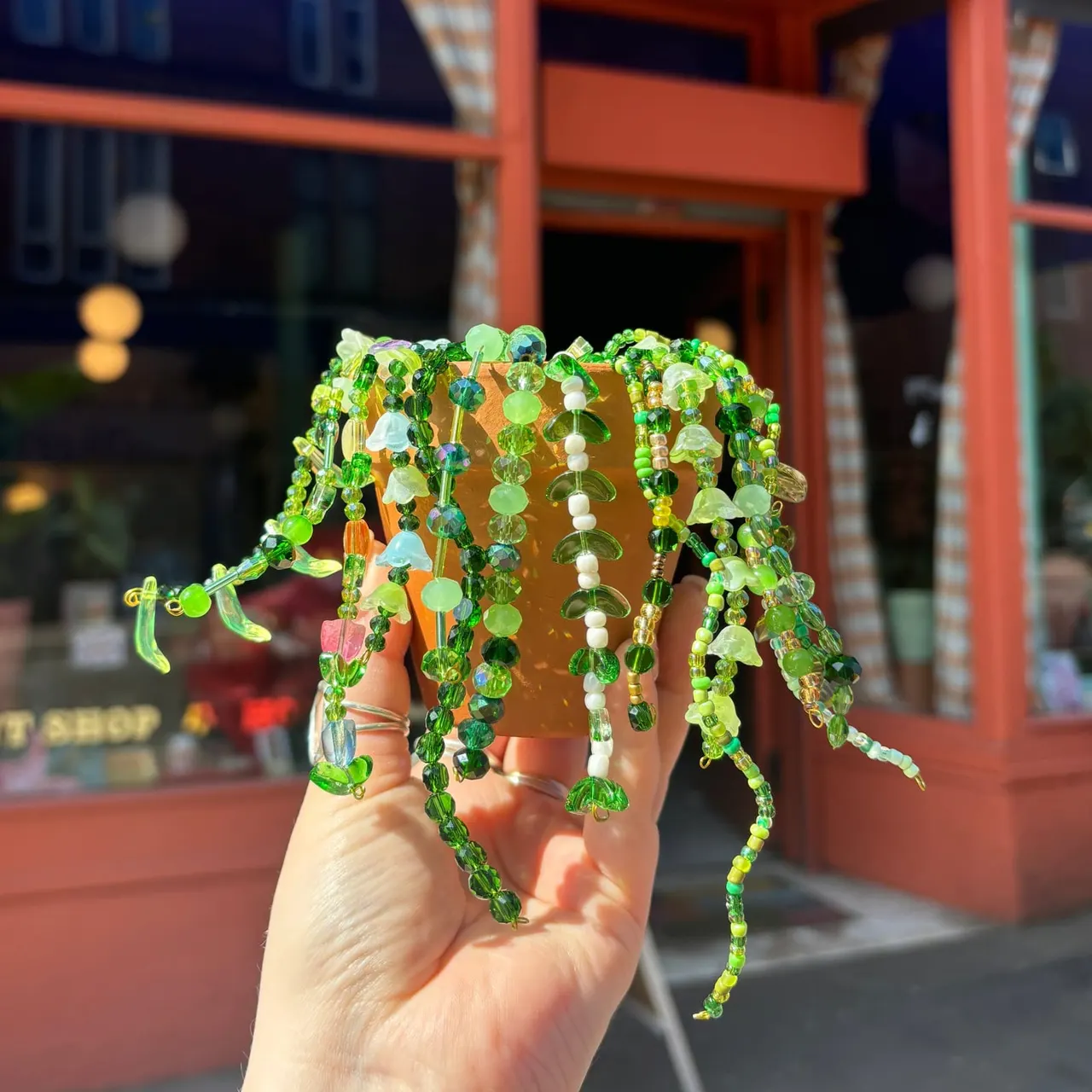 A hand holding a decorative beaded plant pot with cascading green and white beaded strands in front of a red-painted storefront.