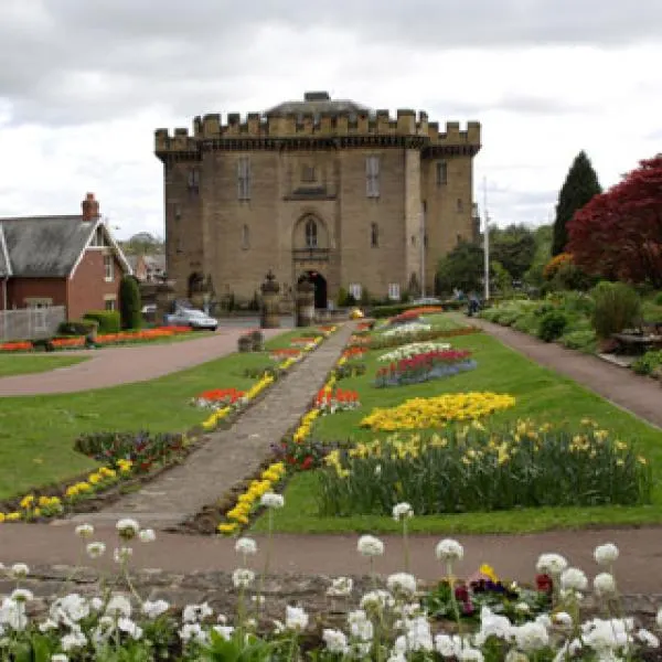 Stone castle with crenellated tower surrounded by formal gardens with colourful flower beds and manicured lawns.