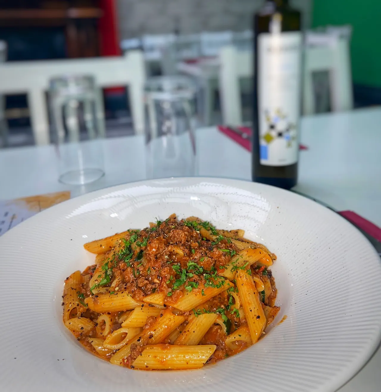Penne pasta with meat ragù sauce and fresh parsley on a white plate at a restaurant table.