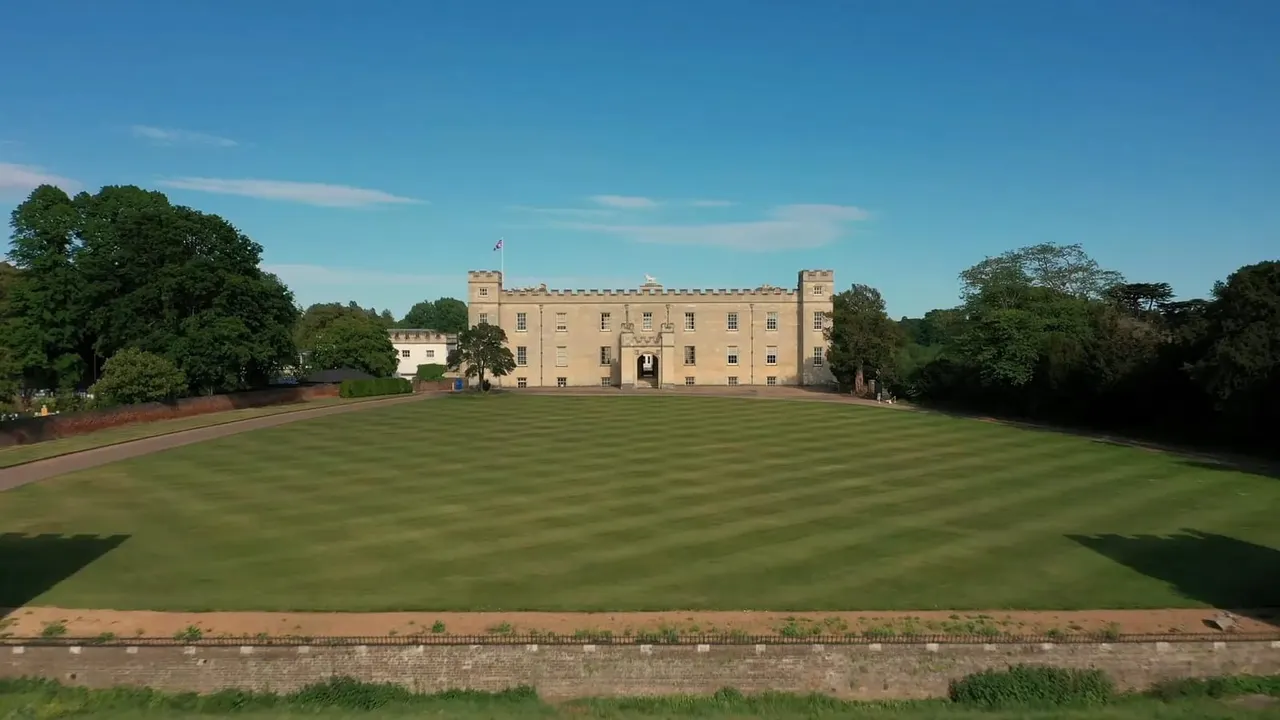 A grand stone castle with manicured lawn and formal gardens stretching across the grounds under clear blue sky.