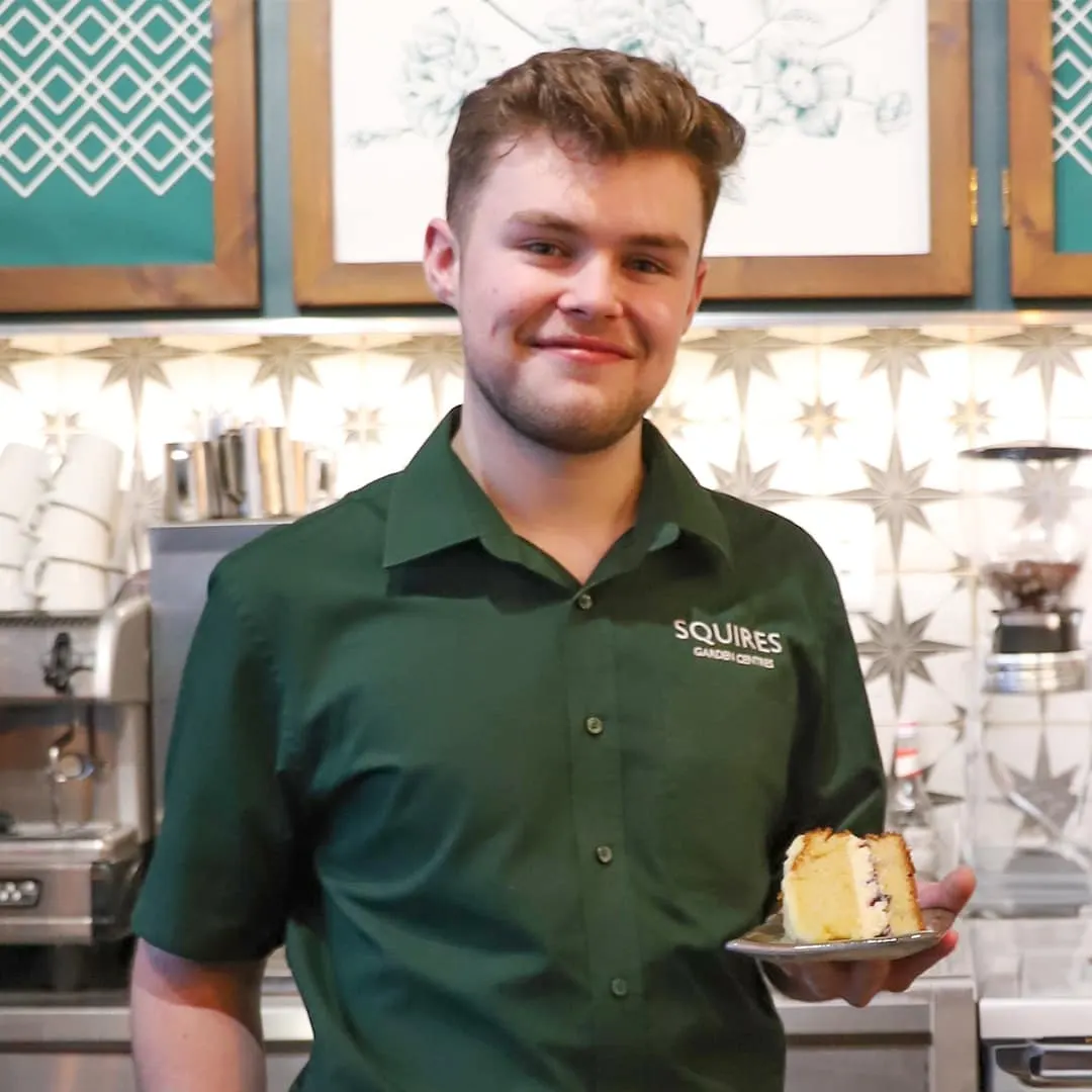 Staff member in green Squires uniform holding a slice of cake in a garden centre café.