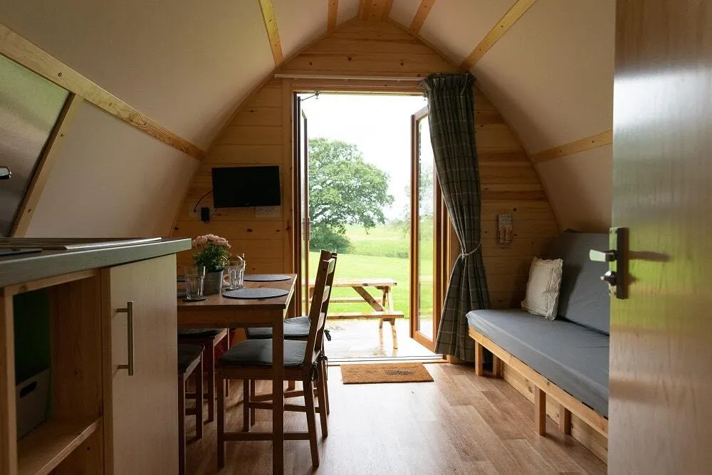 Interior of a wooden glamping pod with open doors revealing countryside view, featuring kitchen area, dining table and bed.