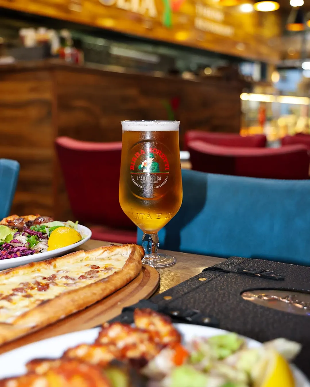 Glass of lager beer and plates of pizza and salad on a restaurant table with colourful booth seating behind.