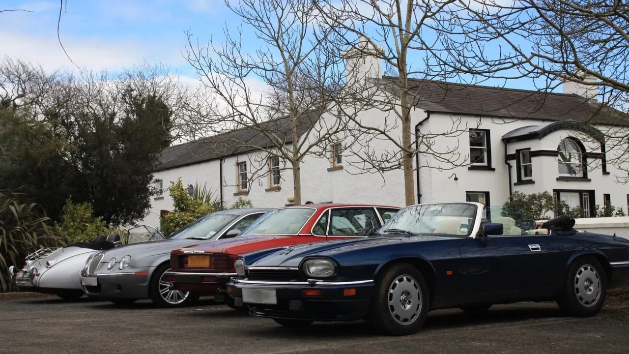 A row of classic and vintage cars parked in front of a white-painted country house with bare trees.