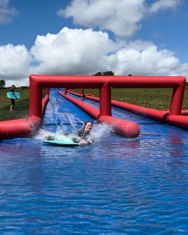Child on a bodyboard splashing through an inflatable water course in an open field.
