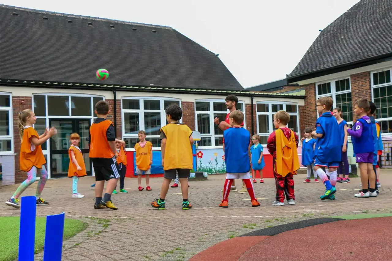 Children in coloured sports bibs participating in an outdoor PE lesson in a school courtyard with brick buildings.