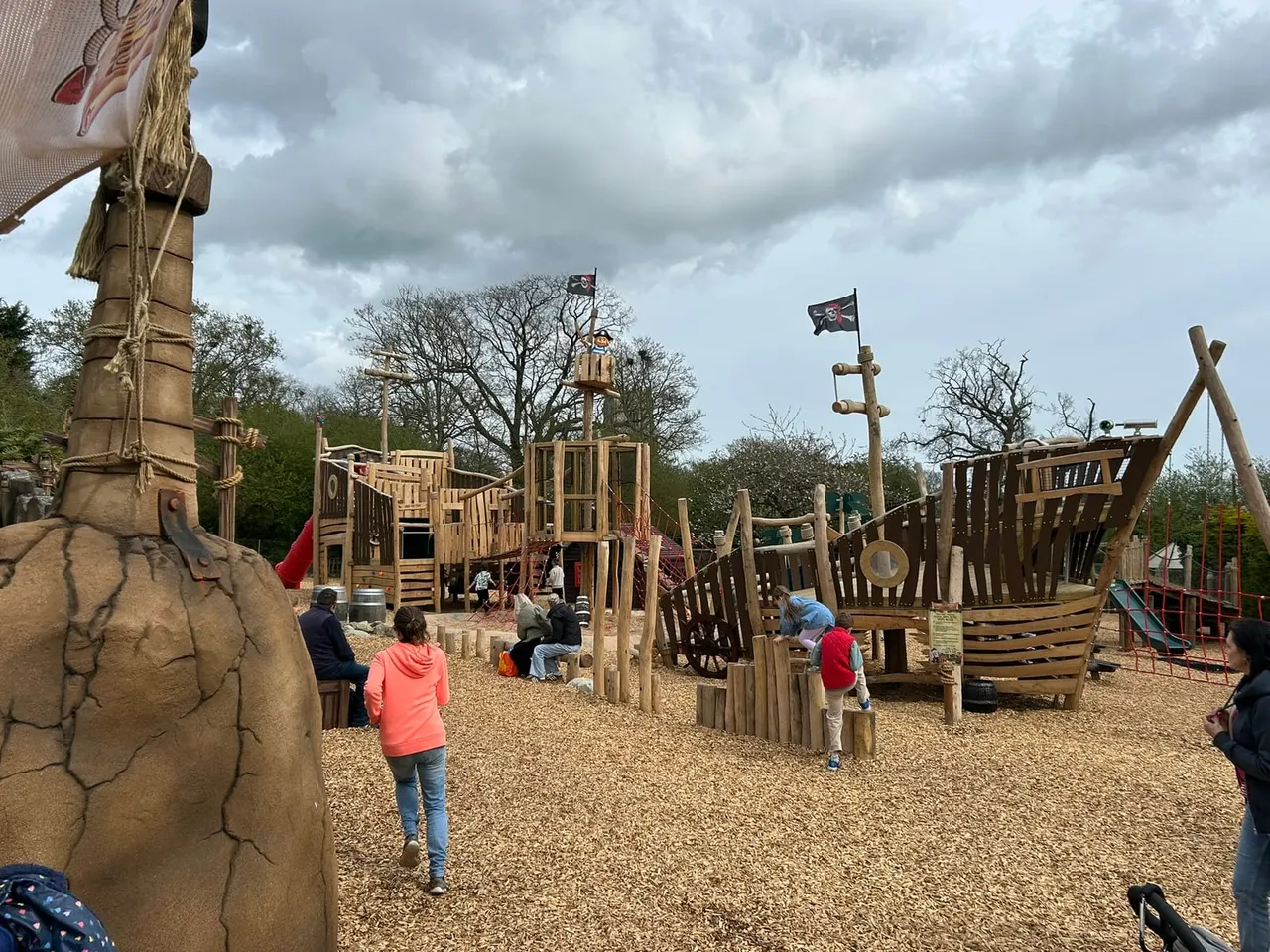 Pirate-themed wooden play structures with children playing on gravel ground beneath cloudy sky.