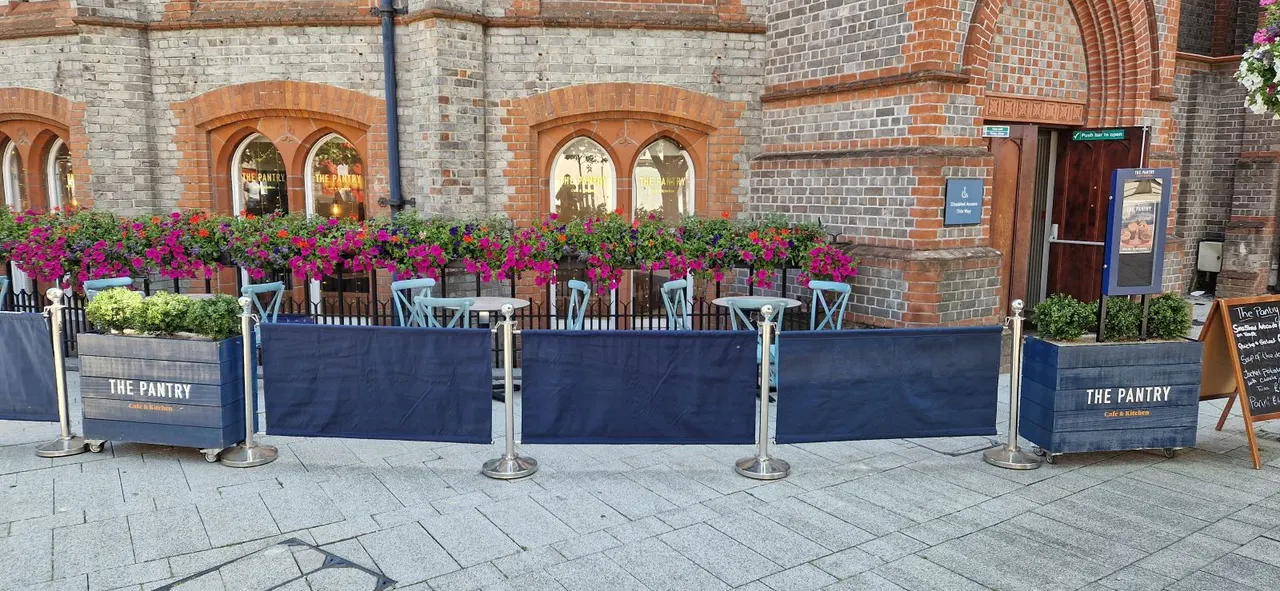 Outdoor seating area with blue barriers and light blue chairs in front of a brick building with arched windows and magenta flowering baskets.
