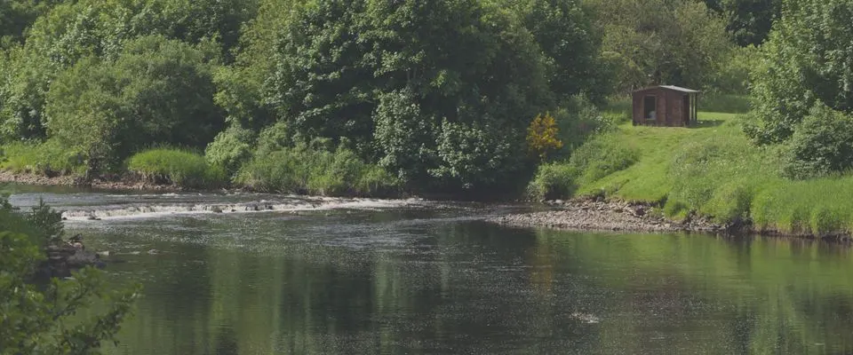 River flowing past lush green banks with a small brick structure visible on the hillside beyond.