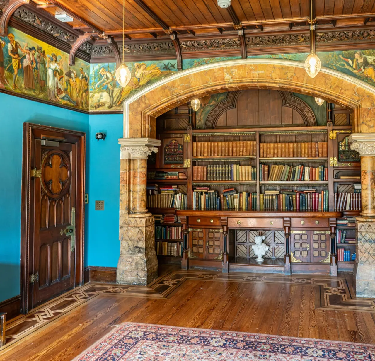 Ornate historic library interior with wooden bookshelves, decorative ceiling panels, turquoise walls and polished parquet flooring.