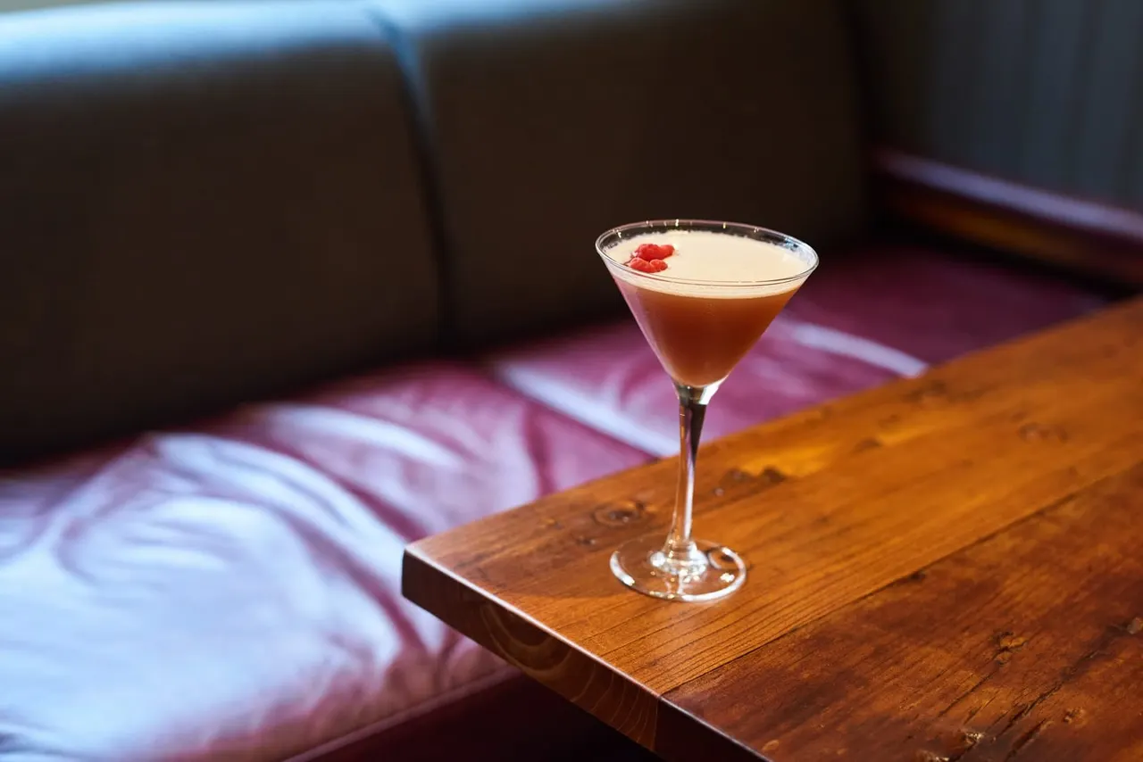 Cocktail with foam top and red garnish on wooden bar counter beside seating.