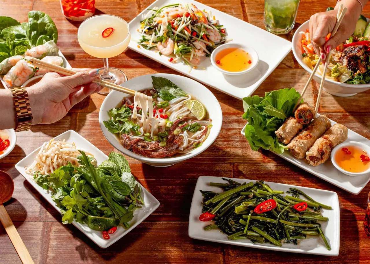 Two diners using chopsticks to eat Vietnamese dishes including pho, spring rolls and stir-fried vegetables at a wooden table.