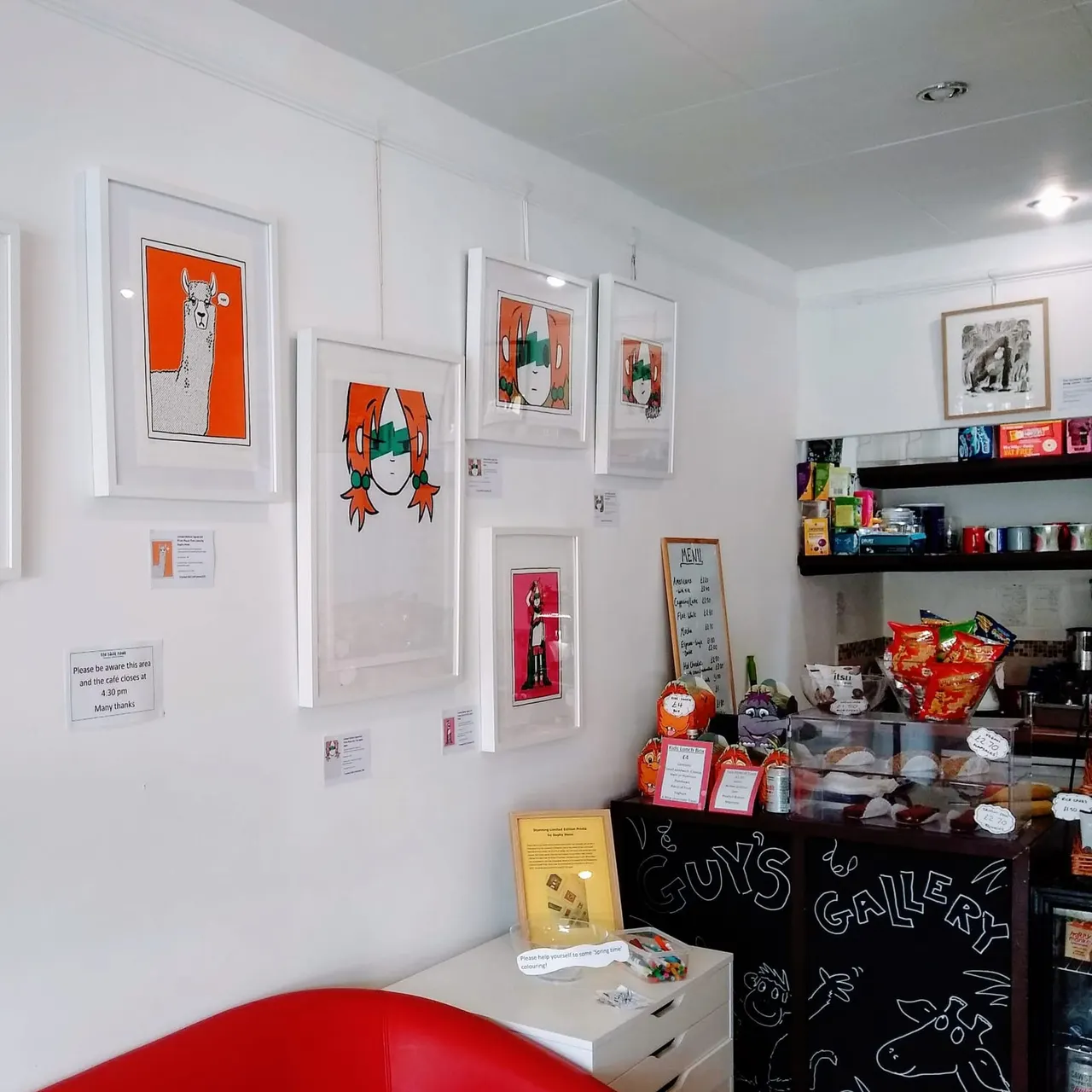 Art gallery wall with colourful pop art prints in white frames, displayed above a counter with merchandise and a black chalkboard sign.