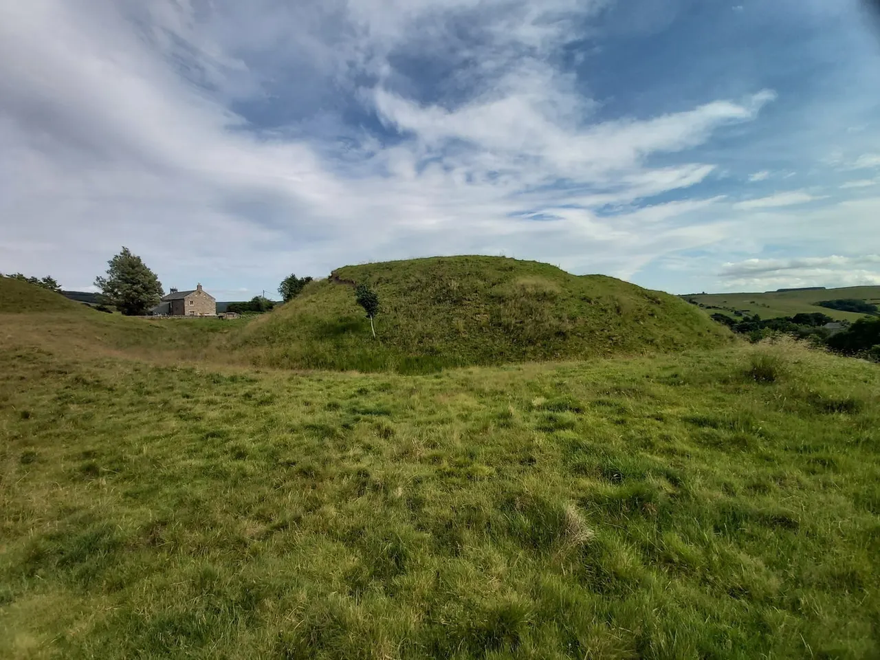 A grass-covered mound with scattered shrubs and trees in a rural landscape, with a stone building visible in the background.