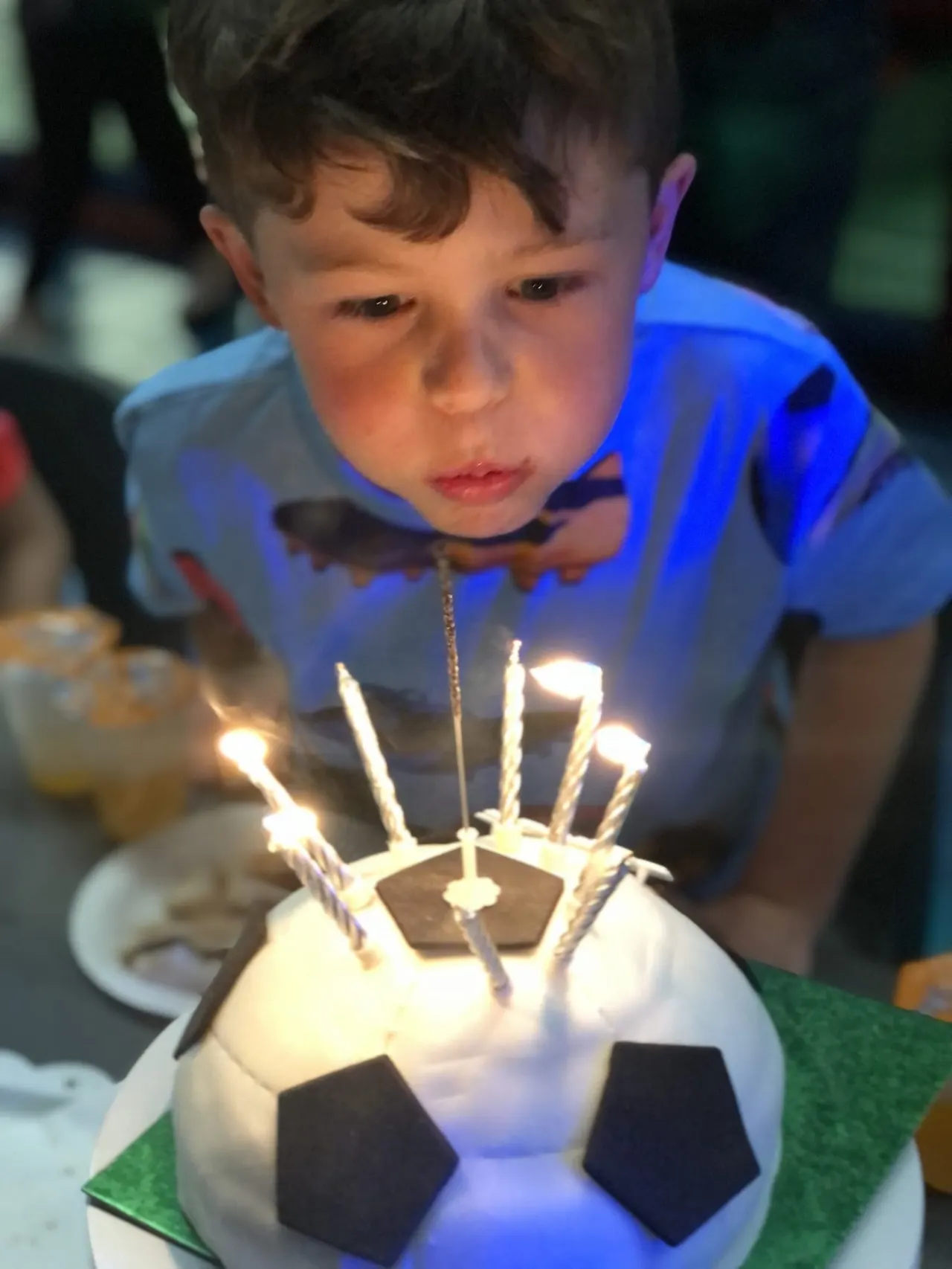 Young child in blue shirt leaning over a football-shaped birthday cake with lit candles.