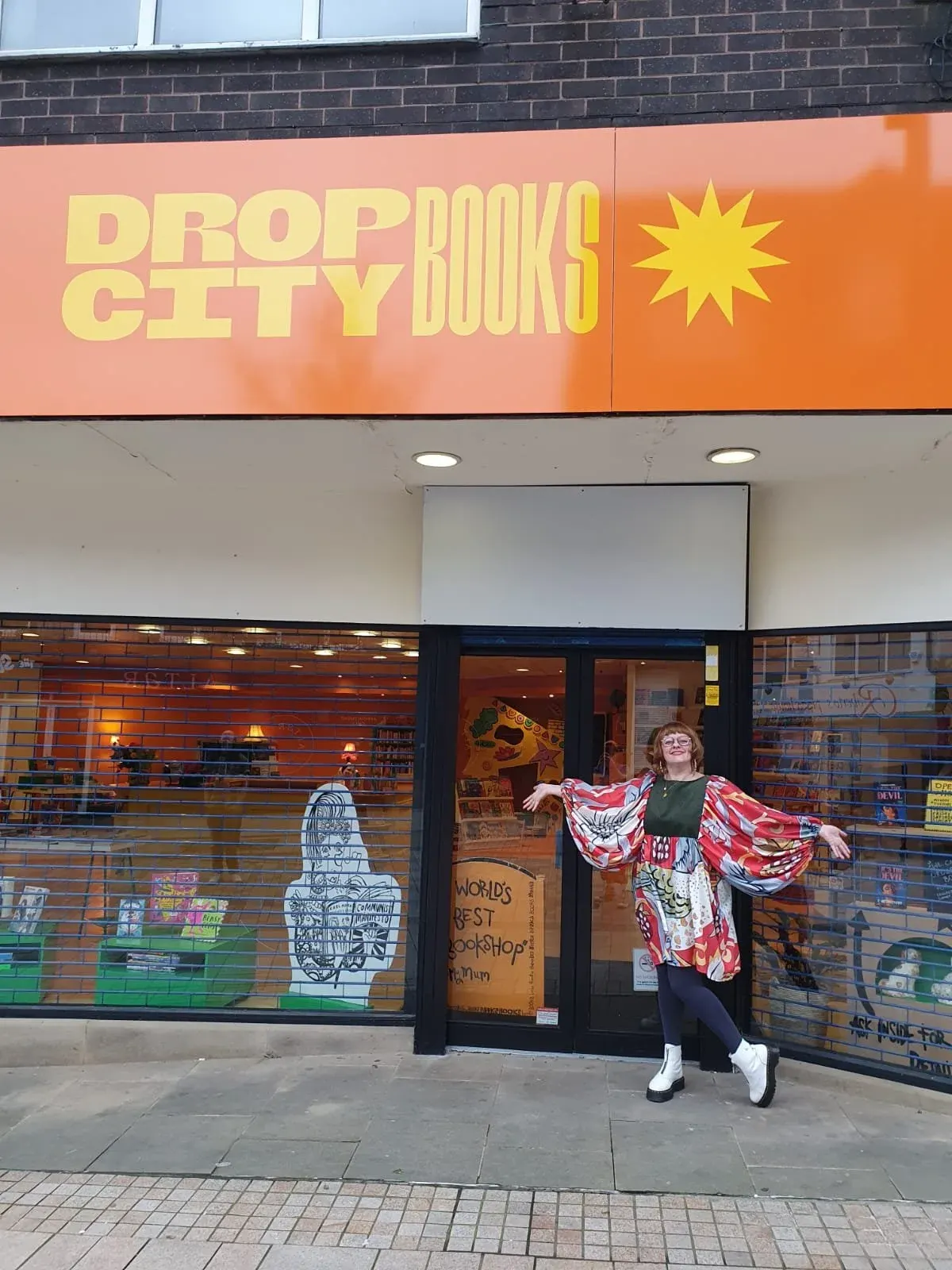 A person posing outside a bookshop entrance with arms outstretched, wearing a colourful patterned jacket.