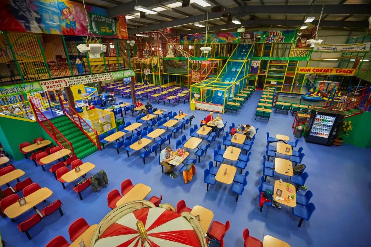 Indoor play centre with colourful climbing frames, slides, seating areas with tables and chairs, and scattered visitors including children.