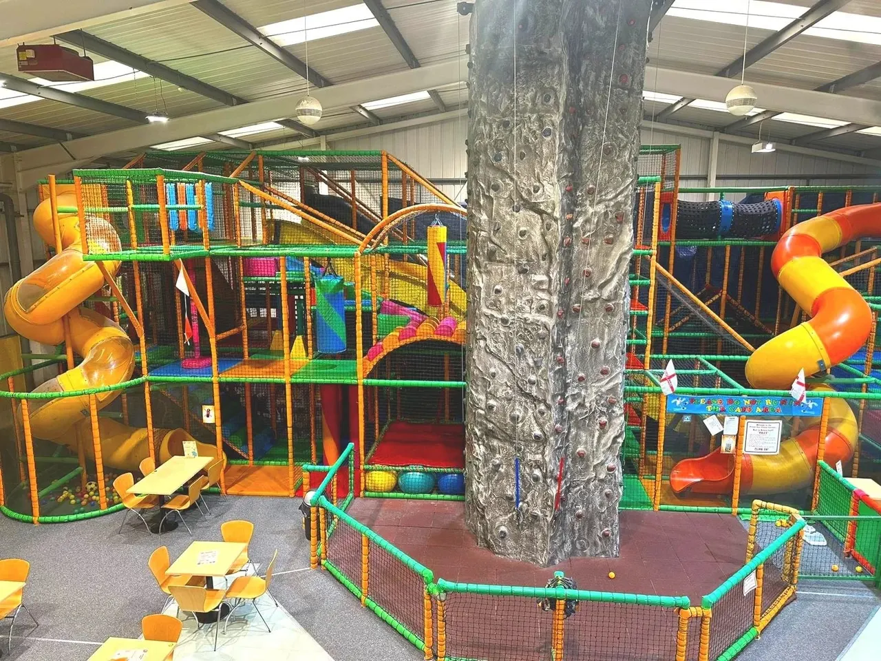 Indoor play structure with slides, tunnels and climbing frames surrounding a support column, with small tables and chairs visible.