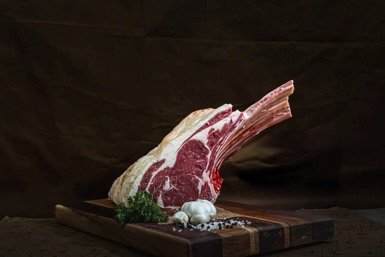Raw tomahawk steak on wooden board with garlic, parsley and sea salt against dark background.