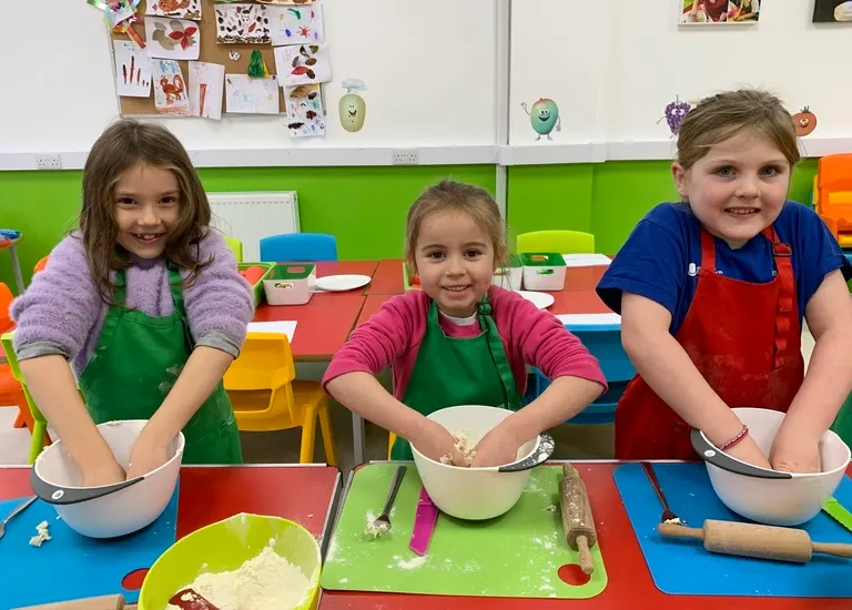 Three children wearing aprons stand at a colourful table with bowls, mixing ingredients whilst cooking together in a bright kitchen classroom.