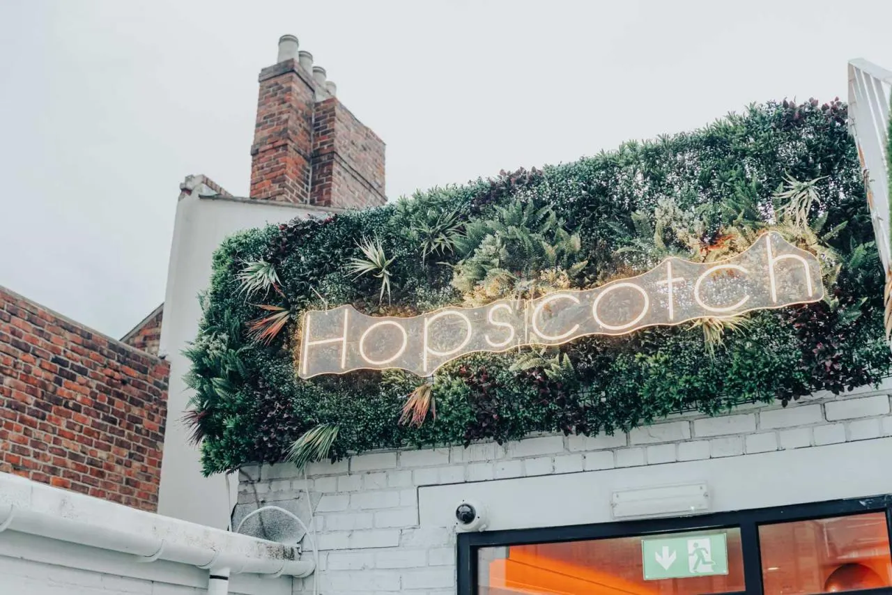 Neon sign reading "Hopscotch" mounted on a green living wall above a white building facade, with a brick chimney visible behind.