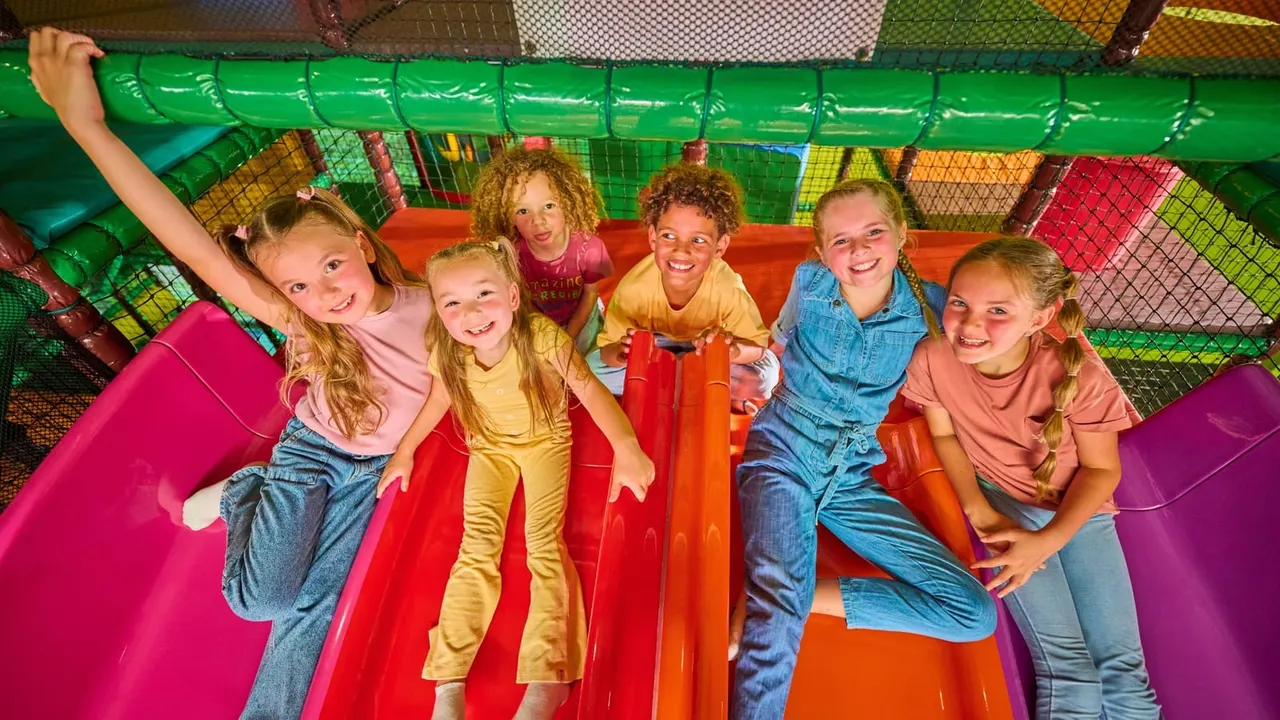 Group of children smiling at camera inside a colourful soft play structure with netting and padded sections.