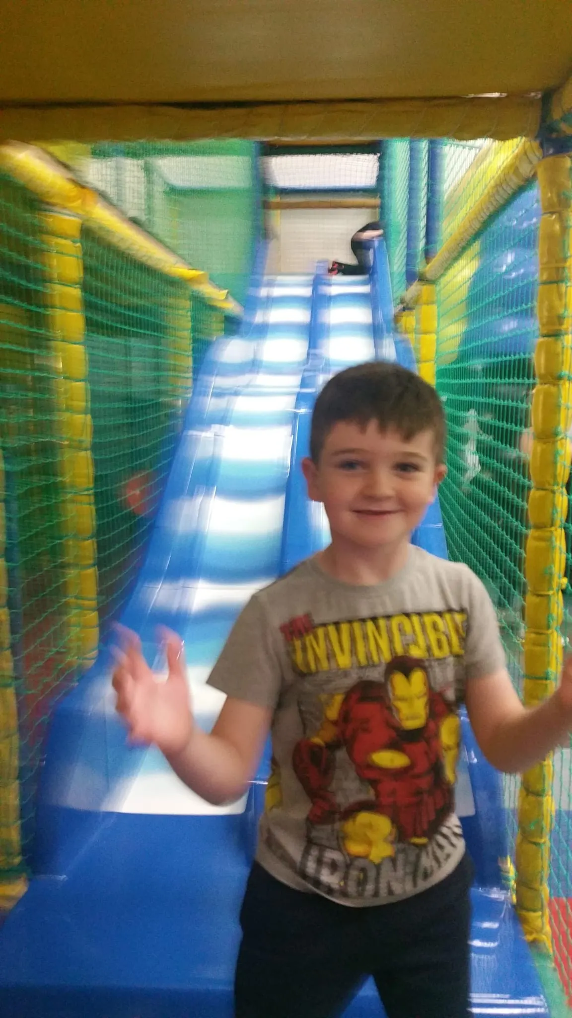 Young child inside a bright yellow and green mesh-walled softplay tunnel with blue padding.