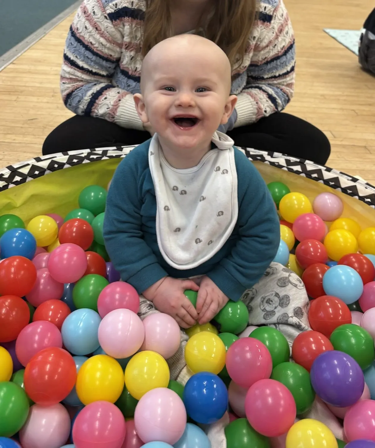 A smiling baby sits in a colourful ball pit whilst being supported by an adult.