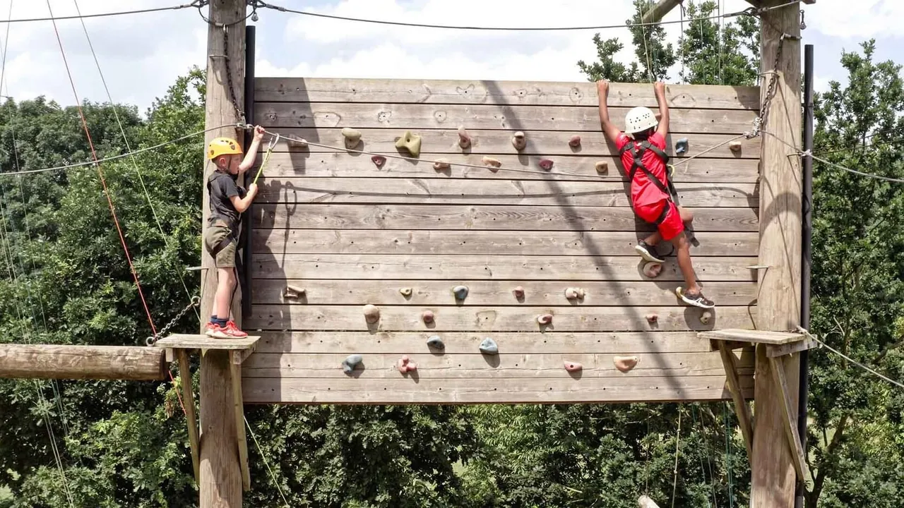 Two people climbing a wooden climbing wall with safety harnesses and helmets in an outdoor adventure park.
