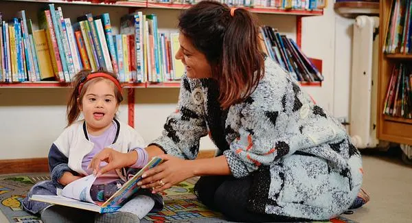 An adult and young child sit together on a colourful mat reading a picture book in front of shelves of books.