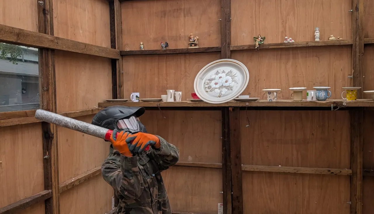 Person in protective gear holding a metal pipe against wooden wall inside rage room.