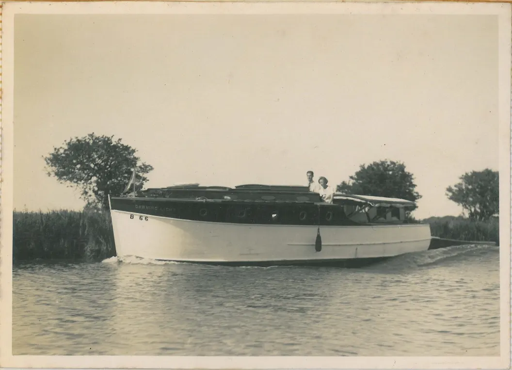 Moored fishing boat with two people aboard on calm water surrounded by reeds and trees.
