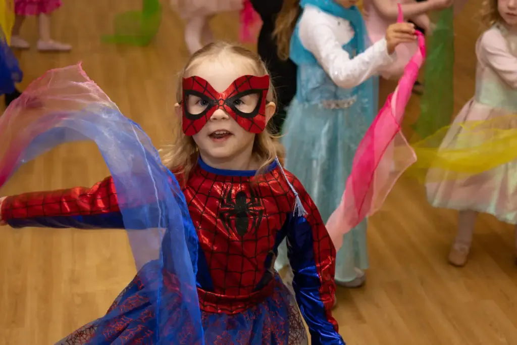 Young child in Spider-Man costume with red mask dancing with other costumed children on a wooden floor indoors.