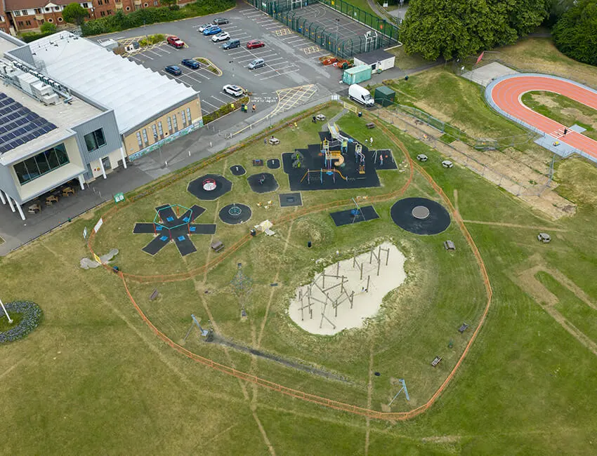 Aerial view of a playground and skatepark with various equipment, trampoline structures, and a sand pit surrounded by grass and pathways.