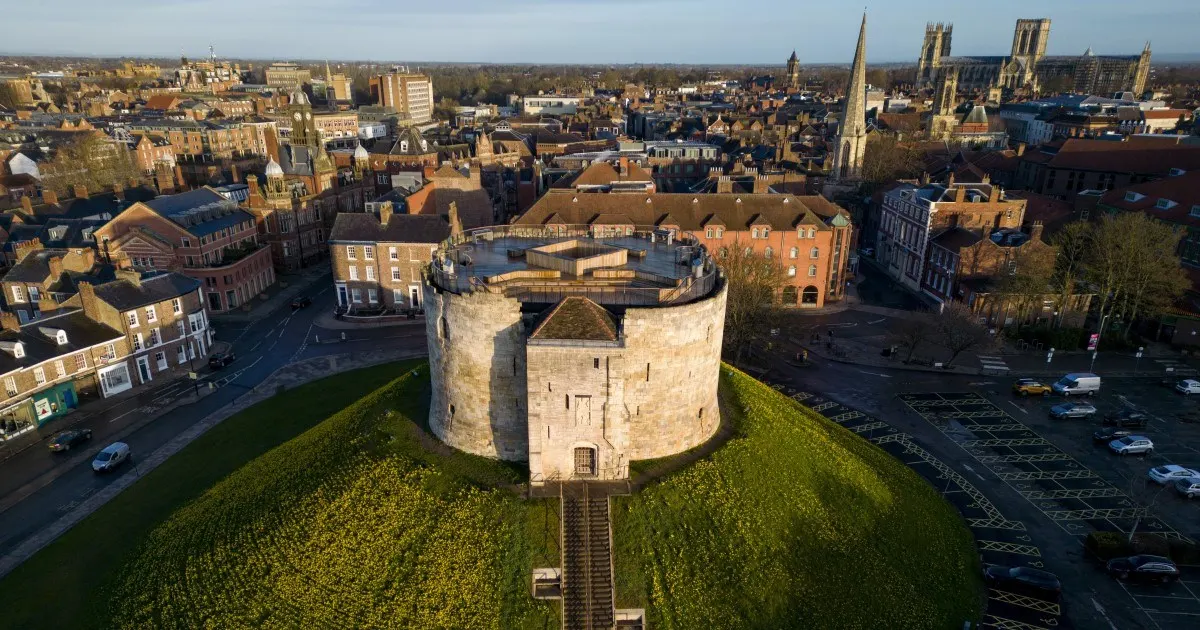Aerial view of a round stone tower surrounded by grass mounds with York cityscape and cathedral in the background.