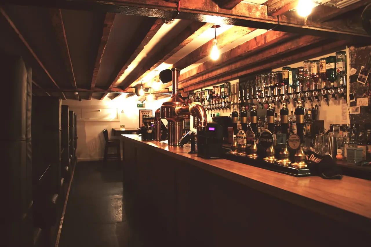 A long bar counter with multiple beer taps and bottles displayed on the wall behind, beneath exposed wooden beams and warm lighting.