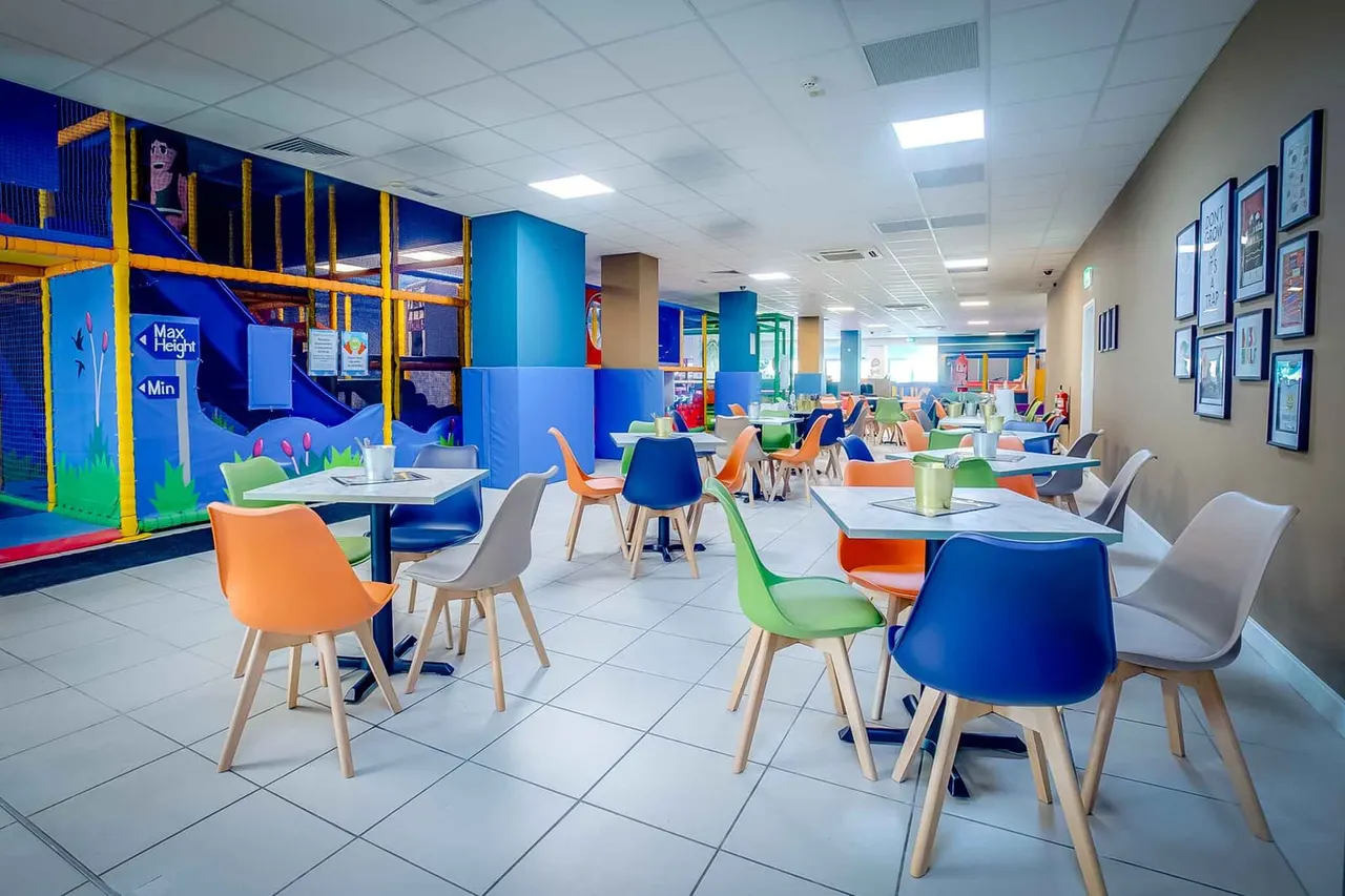 Bright indoor soft play facility with colourful tables and chairs, play structures with yellow and blue frames, and climbing equipment in the background.
