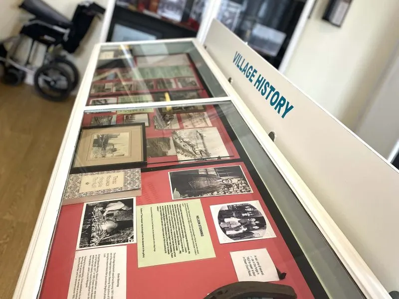 Display case showing historical photographs and documents arranged on red and cream backgrounds with 'Village History' label.