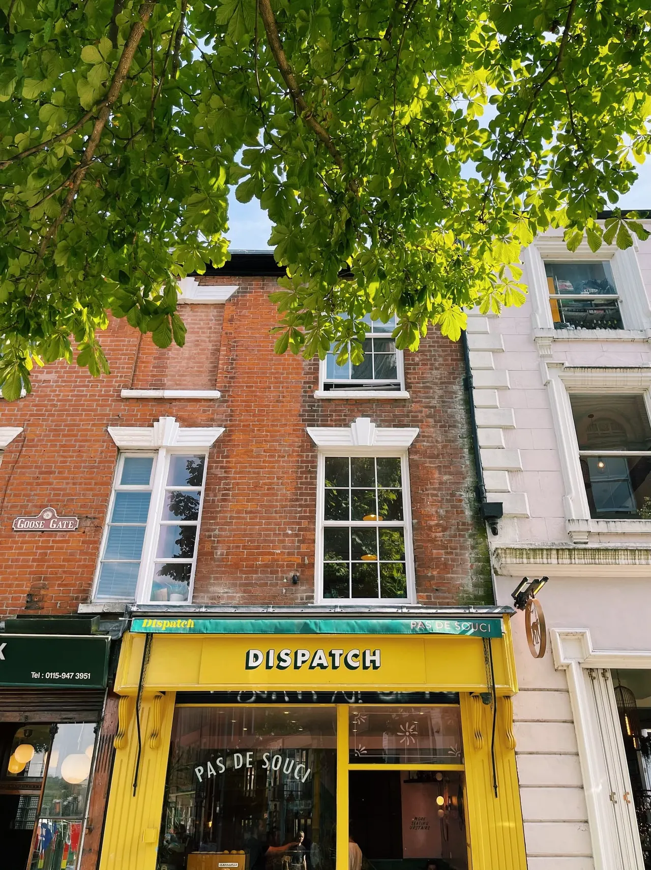 Tree branches with bright green foliage overhang a street frontage with a yellow storefront and red-brick building above.